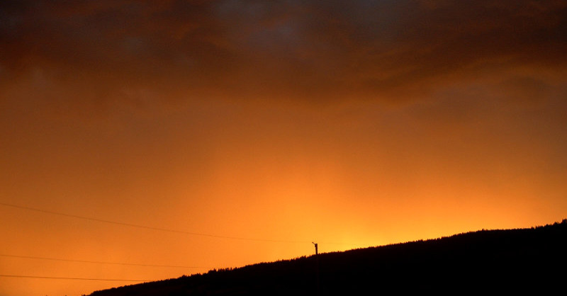 Photograph of an extremely ominous sky: dark orange and with thick black clouds lit up by fire behind a silhouetted ridge.